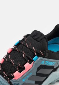 Chaussure de sport noire et grise avec une étiquette Gore-Tex, une tige en mesh, des accents roses et des éléments bleus avec une surface texturée.