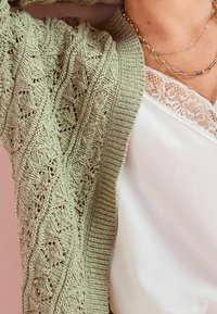 Hellgrüne gestrickte Strickjacke mit filigranem Spitzenmuster und gerippten Kanten, kombiniert mit einem weißen Oberteil mit Spitzenbesatz.