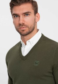Donkergroen v-hals sweater gemaakt van gebreide stof, met een klein geborduurd logo op de borst en een wit overhemd eronder.