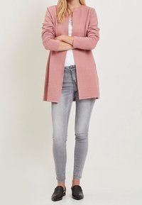 Vaaleanpunainen teksturoitu neuletakki pitkähihaisilla, avonaisella edellä, yhdistettynä istuviin vaaleanharmaisiin farkkuihin ja mustiin loafer-kengiin.