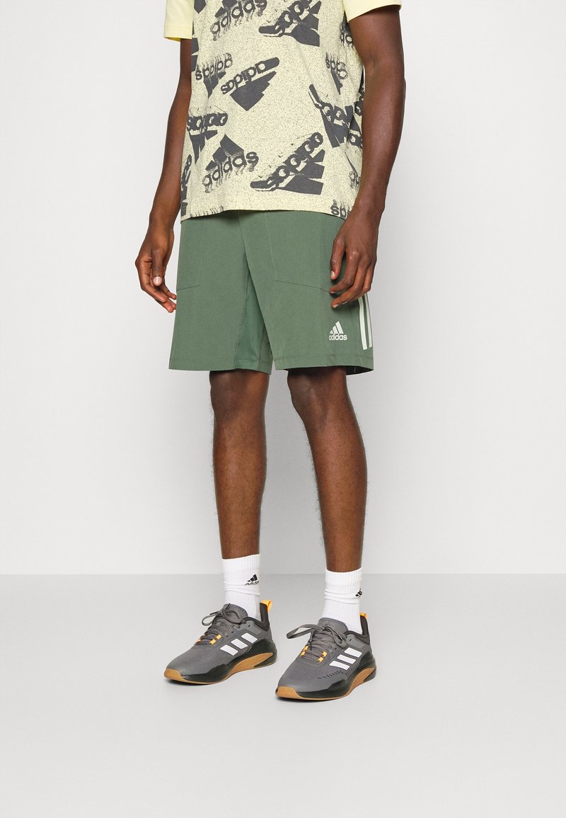 Vihreät urheilushortsit, joissa on kaksi etutaskua ja valkoisia yksityiskohtia, yhdistettynä kuvioituun keltaiseen ja mustaan t-paitaan sekä harmaisiin kenkiin, joissa on oransseja yksityiskohtia.