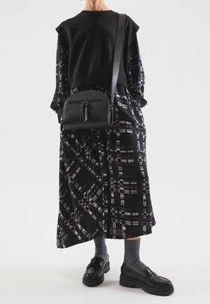 Pull noir sur une robe longue noire à motif. Comprend un sac bandoulière et des mocassins noirs épais avec des chaussettes grises.