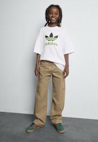 Bílá oversize tričko se zeleno-černým logem Adidas, béžové široké kalhoty a zelené boty s červenými detaily. Hladké textury.