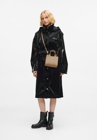 Kobieta w błyszczącej czarnej płaszczu przeciwdeszczowym, czarnych botkach za kostkę i małej, brązowej torebce na ramię z tłoczonym napisem.