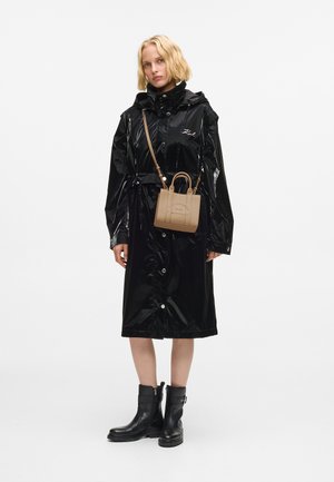 Žena v čiernom lesklom pršiplášti, čiernych členkových čižmách a malej béžovej kabelke cez rameno s embosovaným textom.