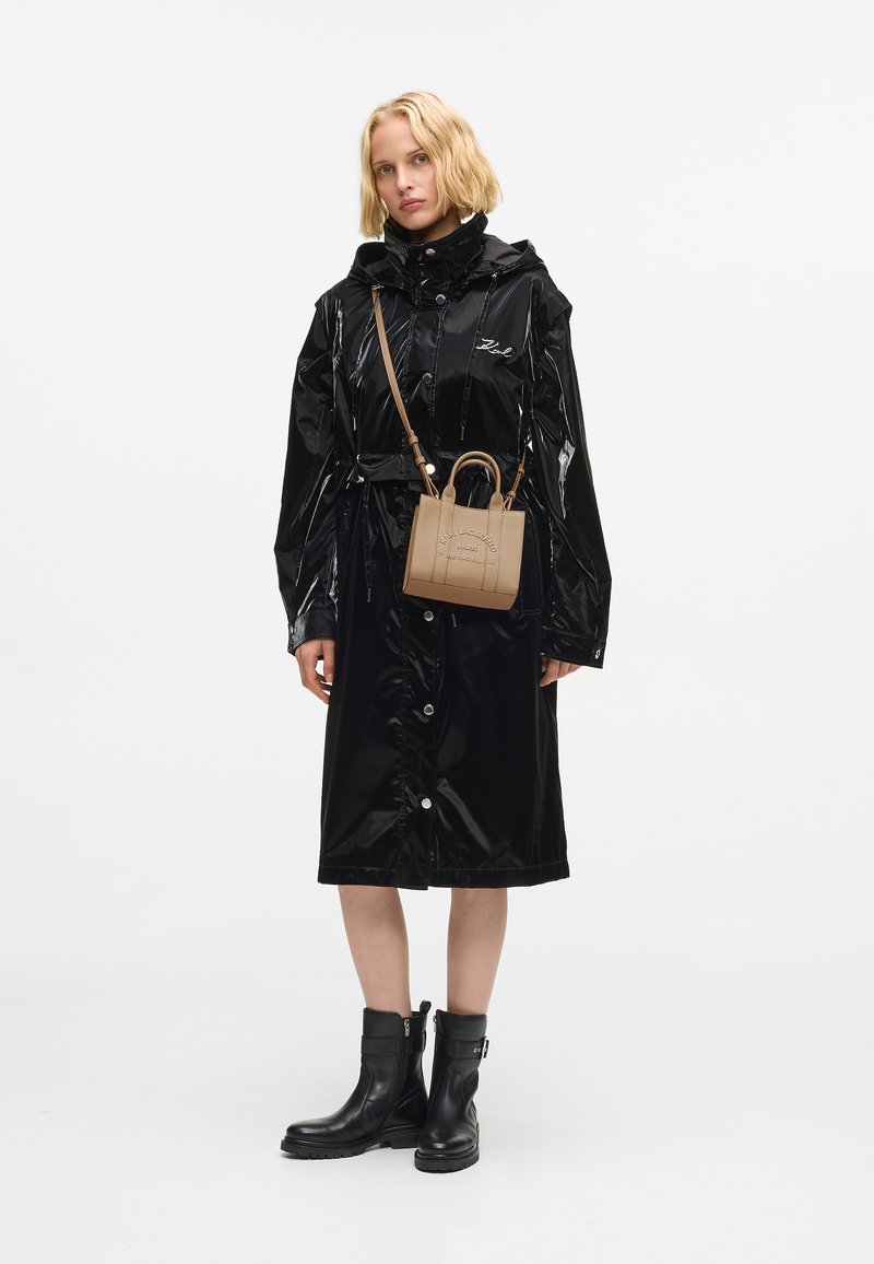 Kobieta w błyszczącej czarnej płaszczu przeciwdeszczowym, czarnych botkach za kostkę i małej, brązowej torebce na ramię z tłoczonym napisem.