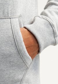 Grijze hoodie met een gestructureerde afwerking, voorzien van een buidelzak en ribbels aan de mouwen. Close-up van een hand die in de zak rust.