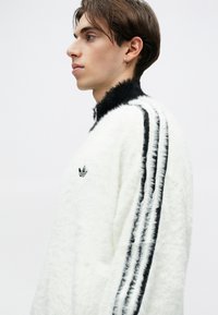 Sudadera blanca esponjosa con un alto cuello negro, presenta tres rayas verticales negras a lo largo de las mangas y un logo bordado.