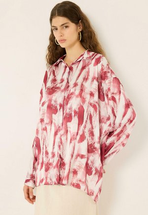 Jonge vrouw met lang golvend haar die gouden oorringen draagt en een oversized wit overhemd met rode abstracte penseelstreken.