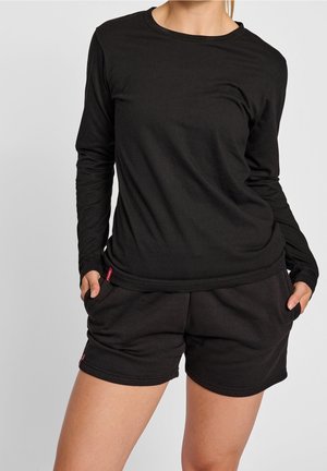 Vrouw die een zwart topje met lange mouwen en zwarte korte broek draagt, met handen in de zakken, staand tegen een effen lichte achtergrond.