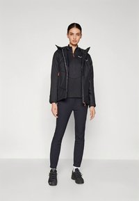 Juoda lauko striukė su užtrauktuku priekyje, derinama su juoda ilgomis rankovėmis ir aptemptomis juodomis kelnėmis. Yra oranžinių akcentų ir faktūruotos medžiagos.