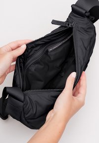 Svart vadderad crossbody-väska med dragkedja, som har en slät inredning, justerbar rem och etikett som visar återvunnet material.