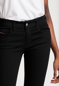 Jeans de mezclilla negra con un diseño ajustado, estilo de cinco bolsillos y un cierre frontal de botón. La tela tiene una textura suave con un ligero estiramiento.