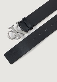Zwarte leren riem met een zilveren metalen CK-logo gesp. Vlotte textuur met gelijkmatig verdeelde gaten langs de band voor aanpassing.