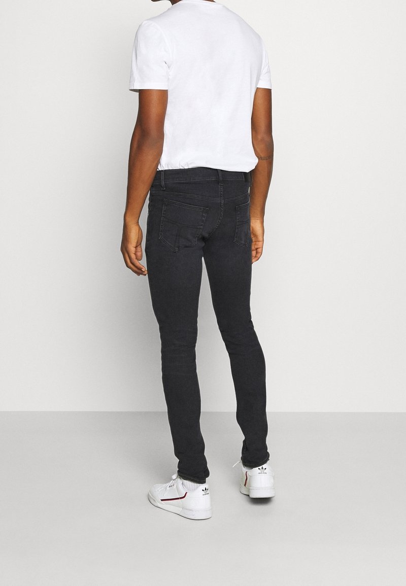 Černé úzké džíny s detaily na zadních kapsách, doplněné o jednoduché bílé tričko a bílé tenisky na neutrálním pozadí.