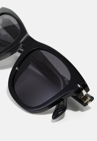 Black oversized sunglasses with dark lenses, glossy finish, prominent square shape, and metal hardware accents on the temples.