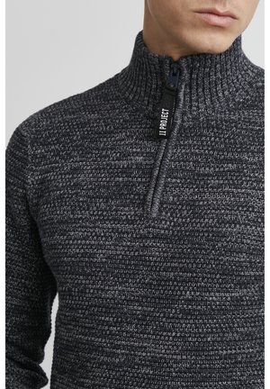 Suéter de punto texturizado en gris oscuro, con un cuello alto y un cierre frontal de media cremallera con un tirador de cremallera de marca.