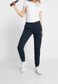 Marine joggerpants van zachte stof, met een elastische tailleband en manchetten, en een logo-detail op de heup. Gecombineerd met een wit poloshirt.