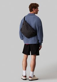 Bolsa de bandolera negra con un patrón, llevada sobre el hombro, que presenta una textura suave y un diseño ligero. El modelo posa con un atuendo casual.