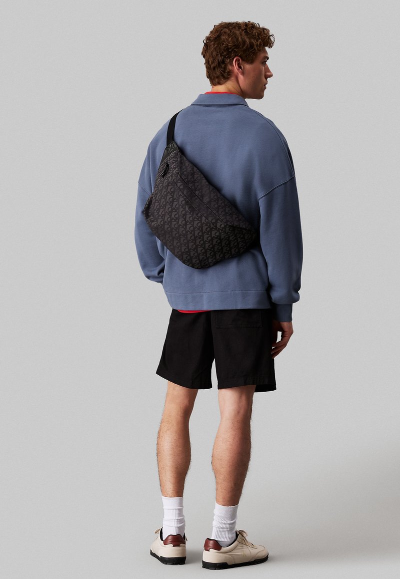 Bolsa de bandolera negra con un patrón, llevada sobre el hombro, que presenta una textura suave y un diseño ligero. El modelo posa con un atuendo casual.