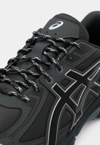 Black athletic shoe with a textured upper, featuring metallic accents and reflective laces. Ergonomic design with a cushioned sole.