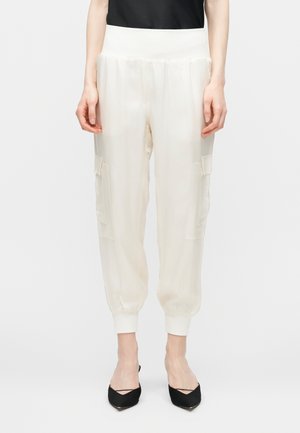 Άτομο που φοράει λευκό παντελόνι jogging cargo με ελαστικές μανσέτες και μέση, σε συνδυασμό με μαύρα slingback παπούτσια με μυτερή μύτη.