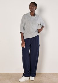Grau, weicher Pullover mit Zopfkragen, kombiniert mit navyblauen, strukturierten, weiten Hosen mit Cargo-Taschen und Knopf-Akzenten. Weiße Sneakers.