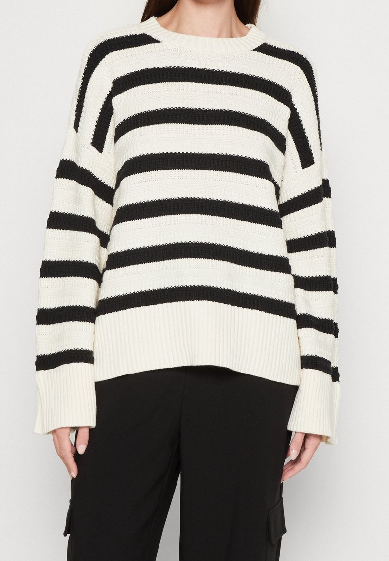 Personne portant un pull en tricot blanc avec de larges rayures horizontales noires et un pantalon noir avec des poches latérales.