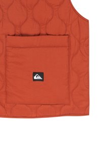 Prošívaná vesta v barvě rezavě oranžové s velkou přední kapsou. Obsahuje malou černou nášivku na kapse. Hladký povrch, bezrukávový design.