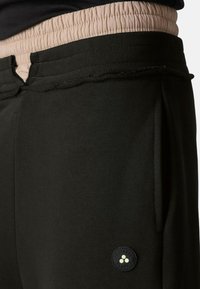 Schwarze Jogginghose mit einem strukturierten Bund in Hellrosa. Verfügt über Seitentaschen und ein rundes Logo-Patch unten links.