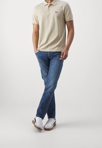 Béžové polo tričko se třemi knoflíky a malým červeným logem, spárované s modrými džíny a bílými teniskami s černými akcenty.