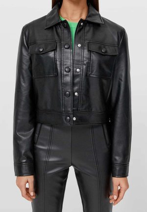 Vrouw draagt een zwarte cropped leren jas met knoopdetails en bijpassende hoge tailler leren broek tegen een effen achtergrond.