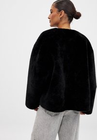 Svart pälsjacka med rund siluett och överdimensionerad form, med en mjuk textur. Bärs med ljusgrå högmidjade jeans.