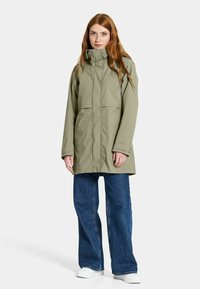 Oliivinvihreä parka, jossa on korkea kaulus, kaksinkertaiset rintataskut ja pitkät hihat, yhdistettynä leveälahkeisiin sinisiin farkkuihin ja valkoisiin lenkkikengiin.