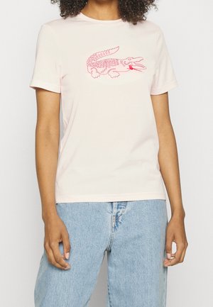 Woman wearing a light pink t-shirt with a red outlined crocodile logo paired with light blue denim jeans against a plain background.