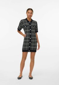Robe en tricot noire avec un motif à carreaux blancs, manches courtes, fermeture à boutons et deux poches poitrine, mettant en valeur des textures contrastées.