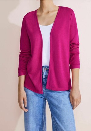 Žena v jasně růžovém cardiganu s otevřeným předním dílem přes bílý top a světle modré džíny, stojící před neutrálním pozadím.