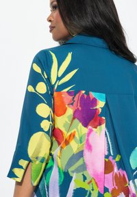 Blusa color verde azulado con un respaldo de patrón floral que presenta colores vibrantes como rosa, amarillo y verde. Textura suave y corte relajado.
