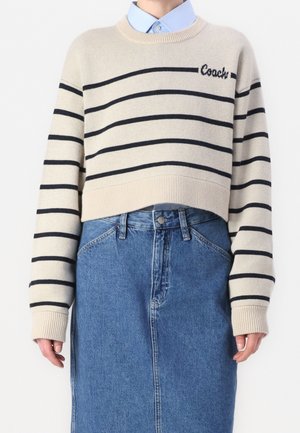 Beige cropped trui met zwarte horizontale strepen en de tekst "Coach", gedragen over een blauw overhemd met kraag en gecombineerd met een blauwe denim rok met hoge taille.