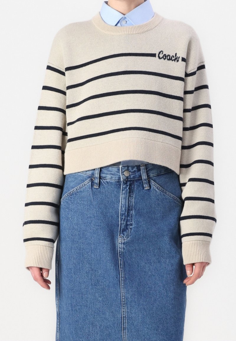 Pull court beige avec des rayures horizontales noires et le texte « Coach », porté sur une chemise bleue à col et associé à une jupe en jean bleue taille haute.