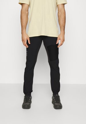 Pantaloni sportivi neri con cintura elastica, gambe affusolate, toppe sulle ginocchia e tasche laterali. Abbinati a una t-shirt beige e scarpe robuste.