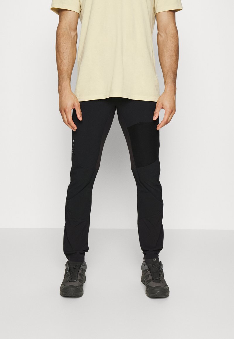 Schwarze Sporthosen mit elastischem Bund, schmal zulaufenden Beinen, Knietaschen und seitlichen Taschen. Kombiniert mit einem beigen T-Shirt und robusten Schuhen.