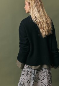 Zwarte gebreide trui met geribbelde zoom en lange mouwen, gedragen over een gelaagde lichtgroene blouse en een doorzichtige rok met een patroon van zwarte vlekken.