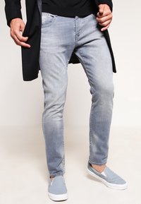 Vaqueros de mezclilla azul claro con corte slim, con un acabado desgastado, diseño de cinco bolsillos y aspecto usado. Combinados con zapatos slip-on de color gris claro.