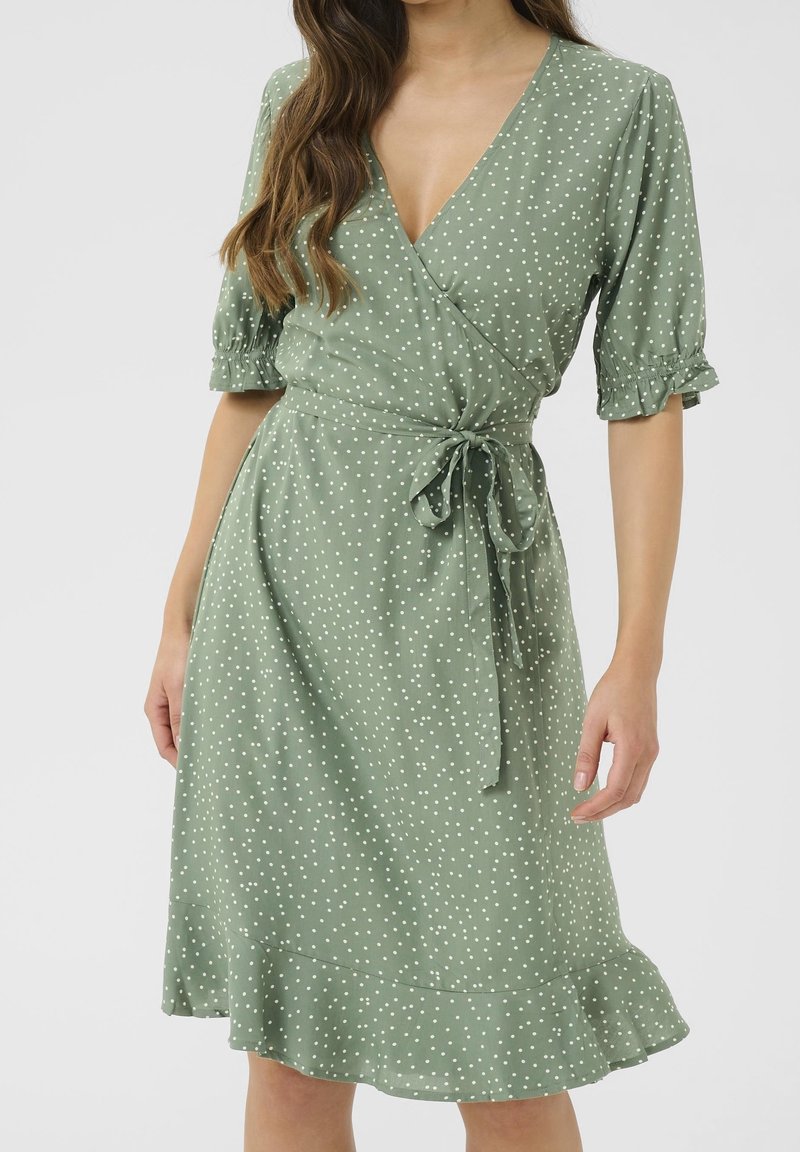 Femme portant une robe portefeuille vert clair à pois blancs, manches courtes bouffantes et un nœud noué à la taille.