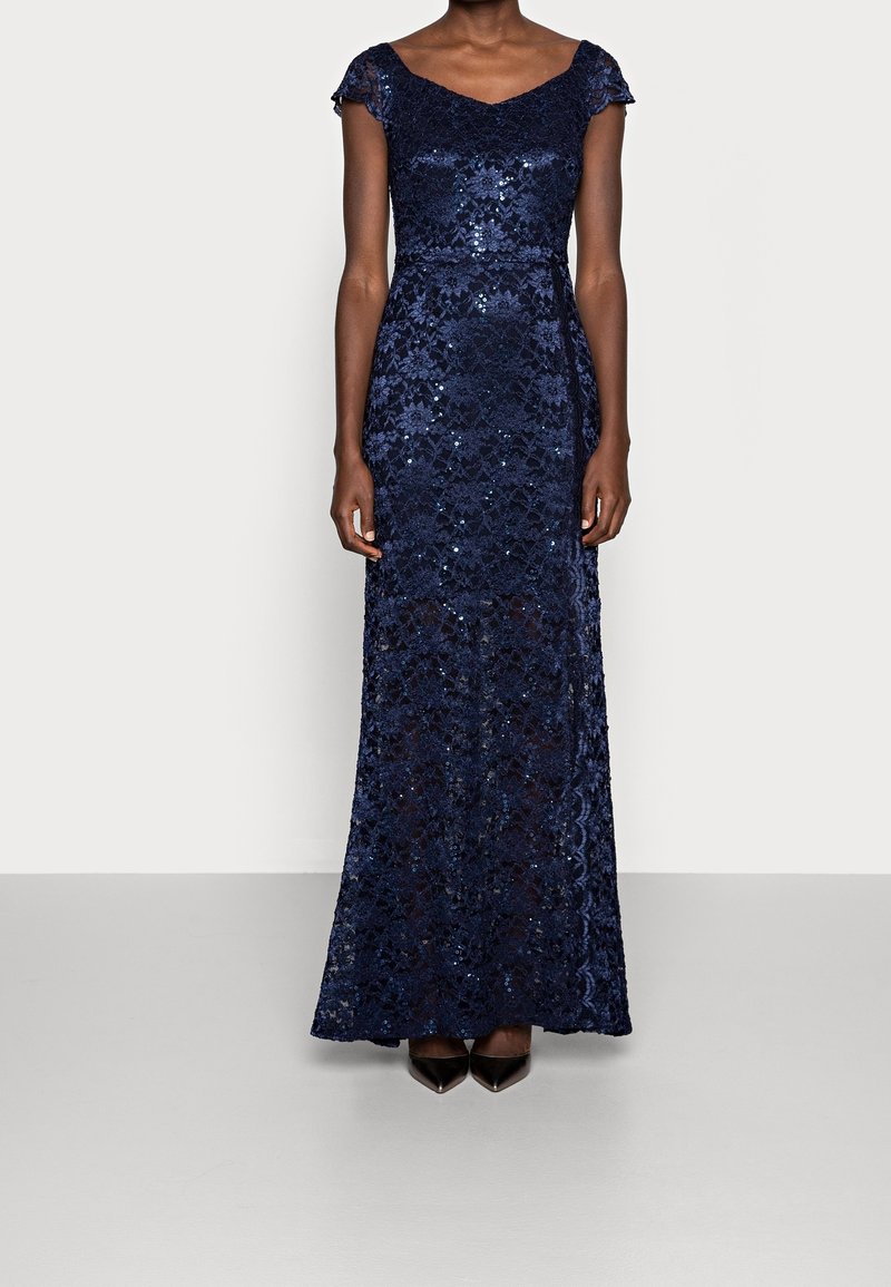 Femme portant une robe longue en dentelle bleu marine avec des manches courtes et des sequins, debout devant un fond uni gris clair.