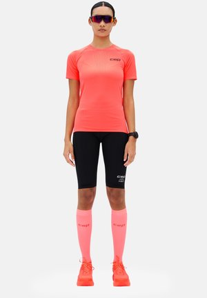 Frau trägt ein korallenrotes Sportshirt, schwarze Radhosen, korallenrote knielange Socken, passende korallenrote Turnschuhe, Sportuhr und Rundum-Sonnenbrille.