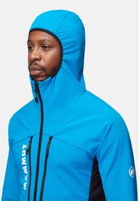 Niebieska kurtka softshellowa z wysokim, dopasowanym kapturem. Posiada czarne akcenty, kieszenie zapinane na zamek z przodu oraz dużą białą grafikę z logo marki na klatce piersiowej.