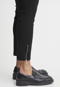 Svarta byxor med en tapered passform, med detaljer som en sidodragkedja vid fållen. Bärs med svarta polerade loafers. Slät textur, skräddarsydd design.