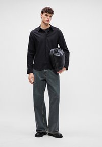 Mustri musta särk, millel on tekstuur, sobitatud lahtiste tumedate teksade ja mustade nahkingadega, hoides musta nööriga kotti.